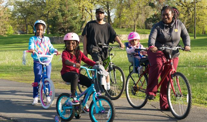 Photo of an African American family on bicycles, dad, mom with a toddler in a seat behind her, and two young girls