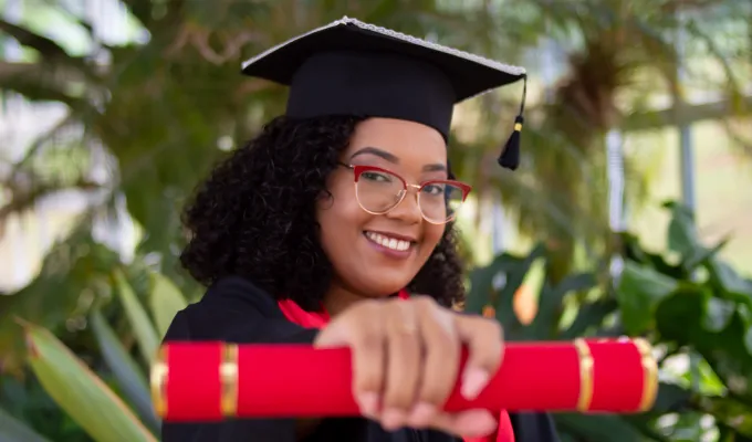 Young Black woman in graduation garb holding our a diploma.