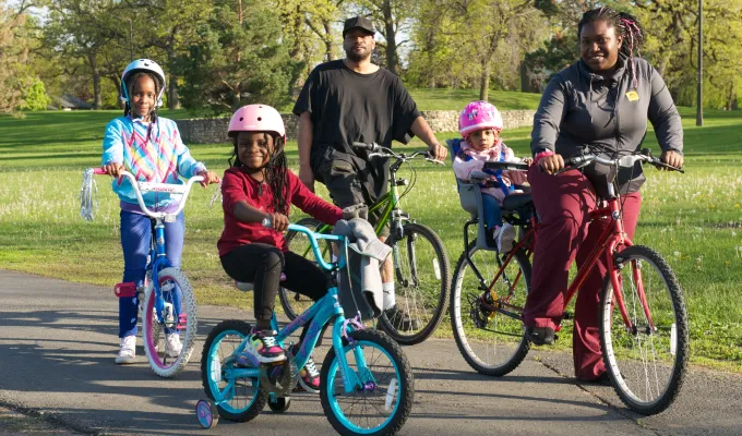 Photo of a family, who are Black, riding bikes--mom, dad, two young girls, and mom has a toddler on a seat behind her.