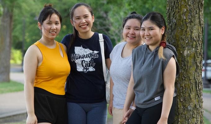 Four young Asian women in a row, smiling at the camera. They are in a park.