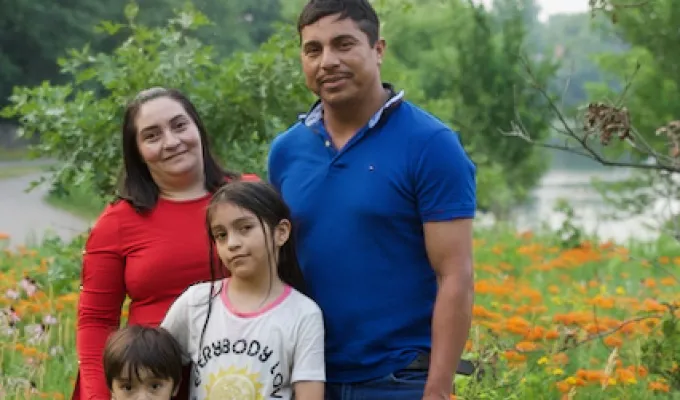 Photo of a Latino Minnesotan family: dad, mom, an elementary-school daughter and a pre-school son. There are orange flowers, trees, and a river in the background.