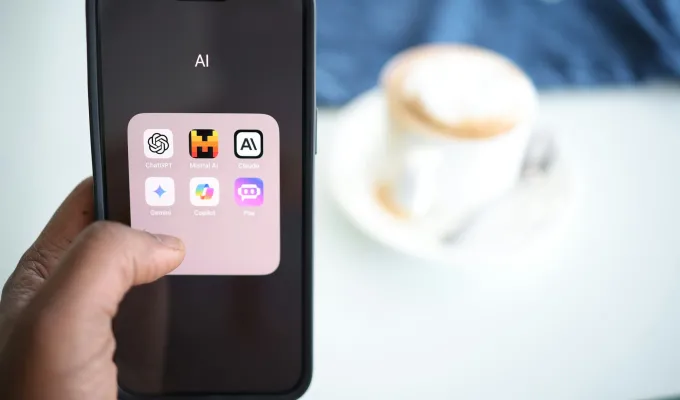 Photo of a hand holding a smart phone showing AI apps on its screen, a cup of coffee in the background. Photo by Solen Feyissa on upsplash