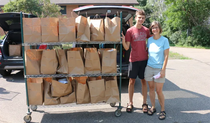 Meals on Wheels volunteers stop to smile at the camera while loading a vehicle with meals. A cart next to them has several grocery bags of food.