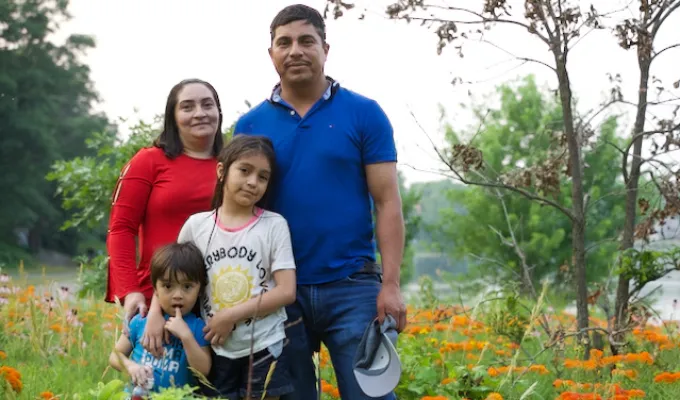 Photo of a Latino Minnesotan family: dad, mom, an elementary-school daughter and a pre-school son. There are orange flowers, trees, and a river in the background.