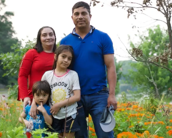 Photo of a Latino Minnesotan family: dad, mom, an elementary-school daughter and a pre-school son. There are orange flowers, trees, and a river in the background.