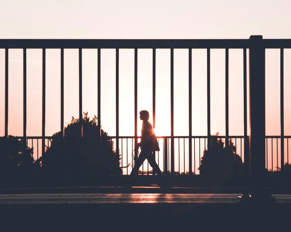 Photo of a man in silhouette walking on a sidewalk against a fence, with a sunset in the background. Photo by Kasper Rasmussen on upsplash.