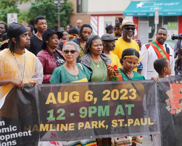 Photo of people in a parade in African dress, holding a sign that advertises and event and is sponsored by African Economic Development Solutions