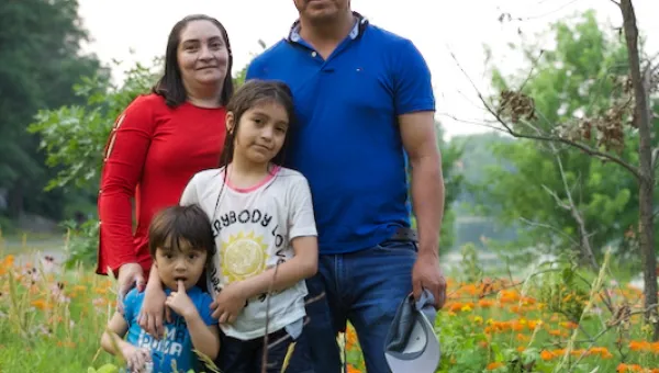 Photo of a Latino Minnesotan family: dad, mom, an elementary-school daughter and a pre-school son. There are orange flowers, trees, and a river in the background.