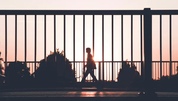 Photo of a man in silhouette walking on a sidewalk against a fence, with a sunset in the background. Photo by Kasper Rasmussen on upsplash.