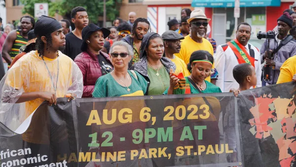 Photo of people in a parade in African dress, holding a sign that advertises and event and is sponsored by African Economic Development Solutions